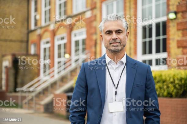 Multiracial male principal in blue jacket, white shirt and name tag around neck. He's standing outside hos school.